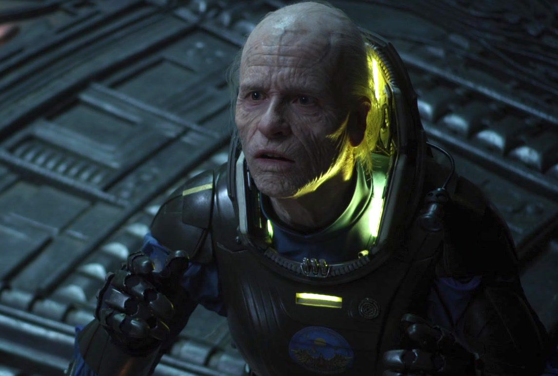 Old Weyland (Guy Pearce en maquillage de vieillesse) dans une combinaison spatiale s'inclinant devant une présence hors écran dans un vaisseau spatial extraterrestre à Prometheus