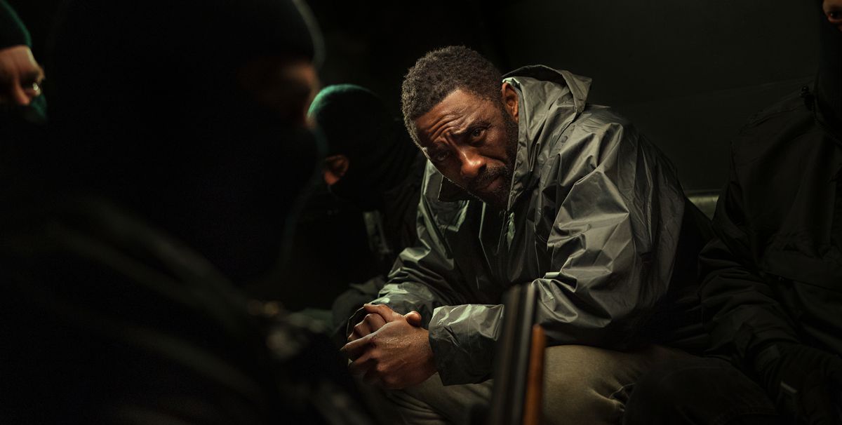 John Luther (Idris Elba) est assis dans une pièce très sombre, les mains jointes devant lui et regarde la caméra, entouré de trois hommes portant des cagoules couvrant le visage, dans Luther: The Fallen Sun