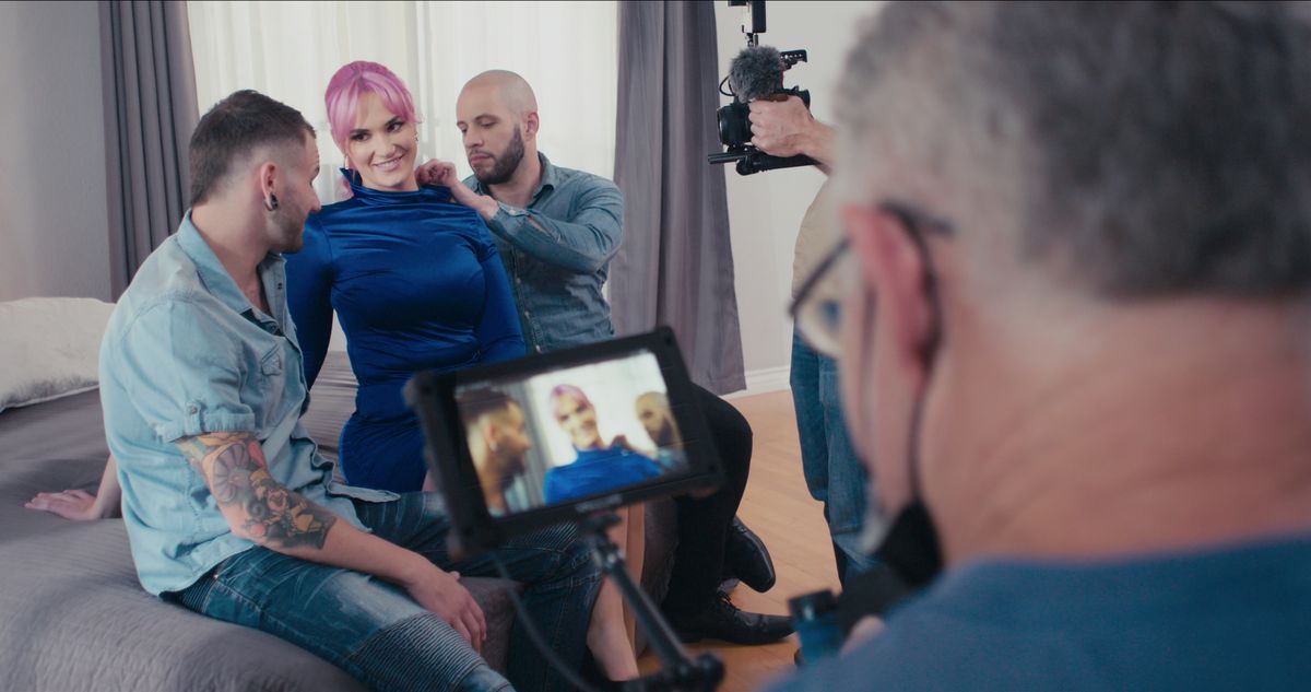 L'interprète de cinéma classée X Siri Dahl, vêtue de soie bleu foncé, est assise sur un lit entre deux hommes alors que les caméramans se concentrent sur eux dans un plan du documentaire Netflix Money Shot: The Pornhub Story