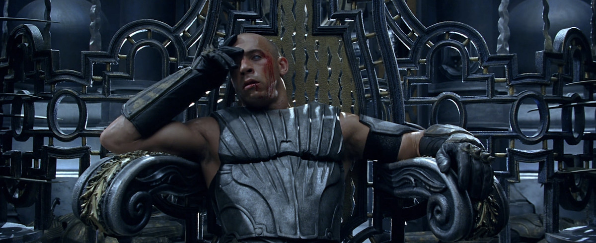 Riddick (Vin Diesel), dégoulinant de sang et portant une armure d'acier grise, est assis sur un trône, la tête appuyée sur sa main dans Les Chroniques de Riddick