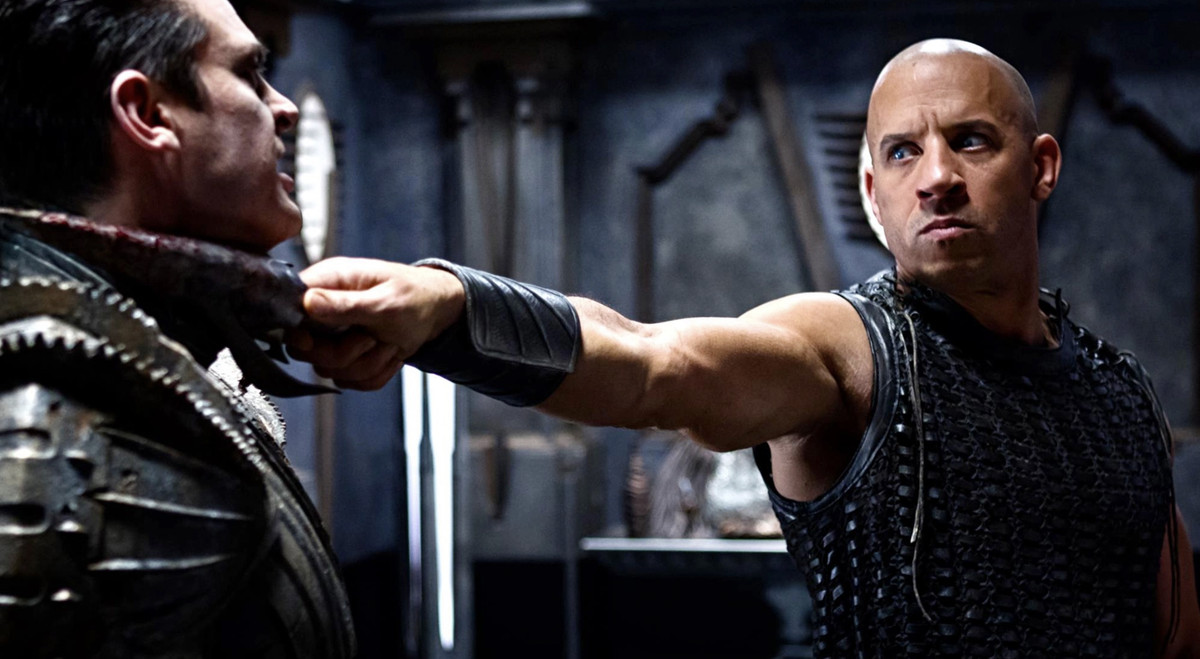 Riddick (Vin Diesel) tient une lame déchiquetée couverte de sang à la gorge d'un homme en armure métallique recouverte de lames de scie rotative dans Riddick de 2013
