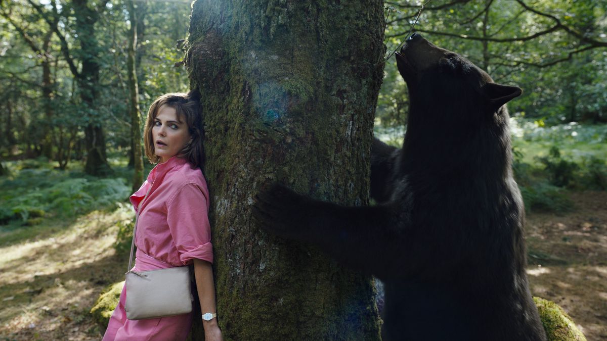 Sari (Keri Russell), une femme à l'air terrifiée vêtue d'une robe rose, se cache derrière un arbre alors qu'un ours bourré de cocaïne renifle de l'autre côté dans Cocaine Bear