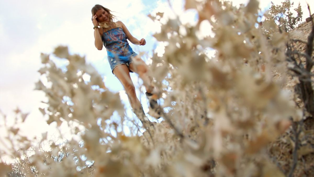 Une jeune femme blonde portant un haut coloré est vue à travers les plantes du désert dans les Outwaters.