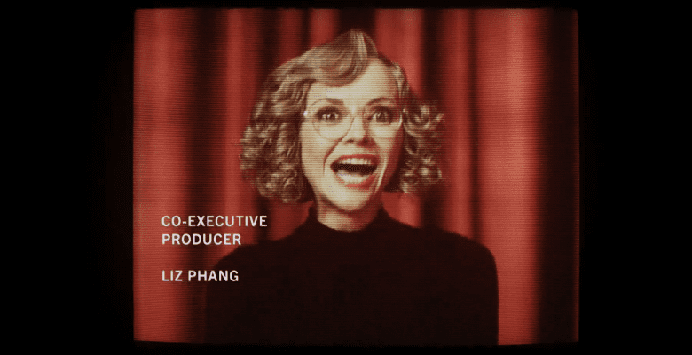 Medium shot of a woman with curly blonde hair and glasses laughing maniacally in front of a red curtain backdrop; still of Christina Ricci in "Yellowjackets"