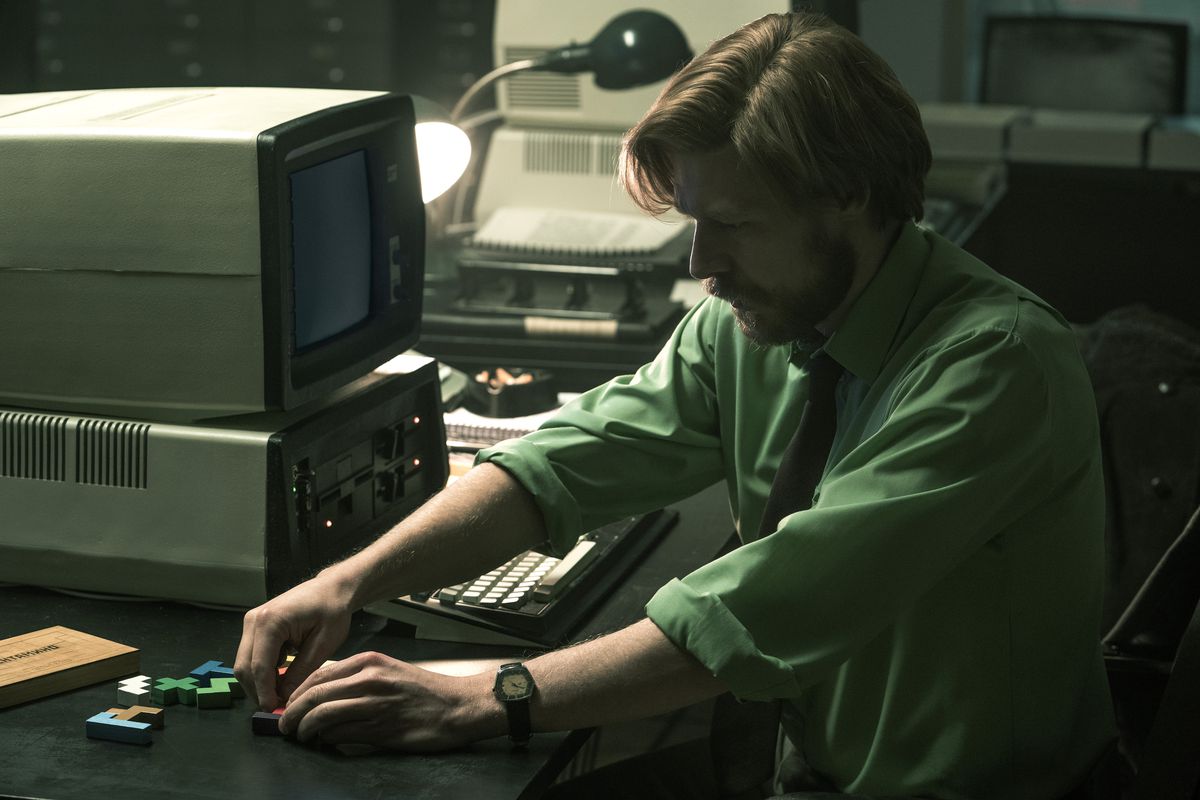 Un jeune homme aux cheveux roux avec une barbe joue avec des pièces de puzzle tetromino sur son bureau à côté d'un ordinateur ancien