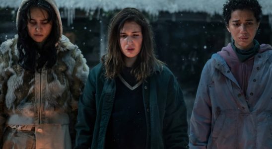Three teen girls in winter outerwear standing outside a snowy cabin and looking down solemnly.