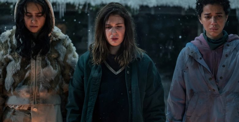 Three teen girls in winter outerwear standing outside a snowy cabin and looking down solemnly.