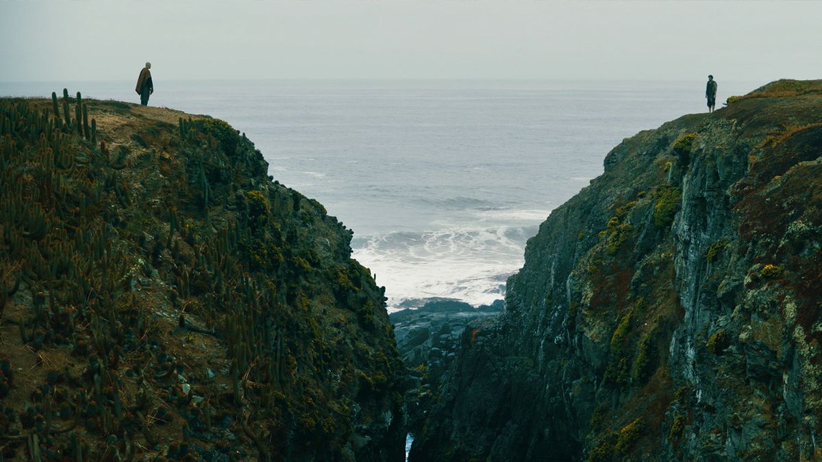 Deux hommes se tiennent au sommet de falaises qui se surplombent, avec un gouffre entre eux et l'océan derrière eux, dans Fist fo the Condor.