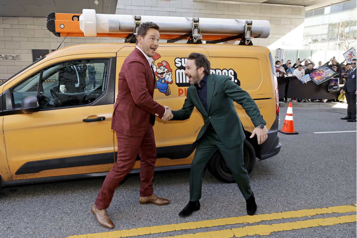 Chris Pratt et Charlie Day se tiennent devant une fourgonnette à thème, lors de la première de Super Mario Bros LA.  Pratt est dans un costume rouge foncé et Day est dans un costume vert foncé, et les deux se serrent la main avec des expressions maladroites sur leurs visages.