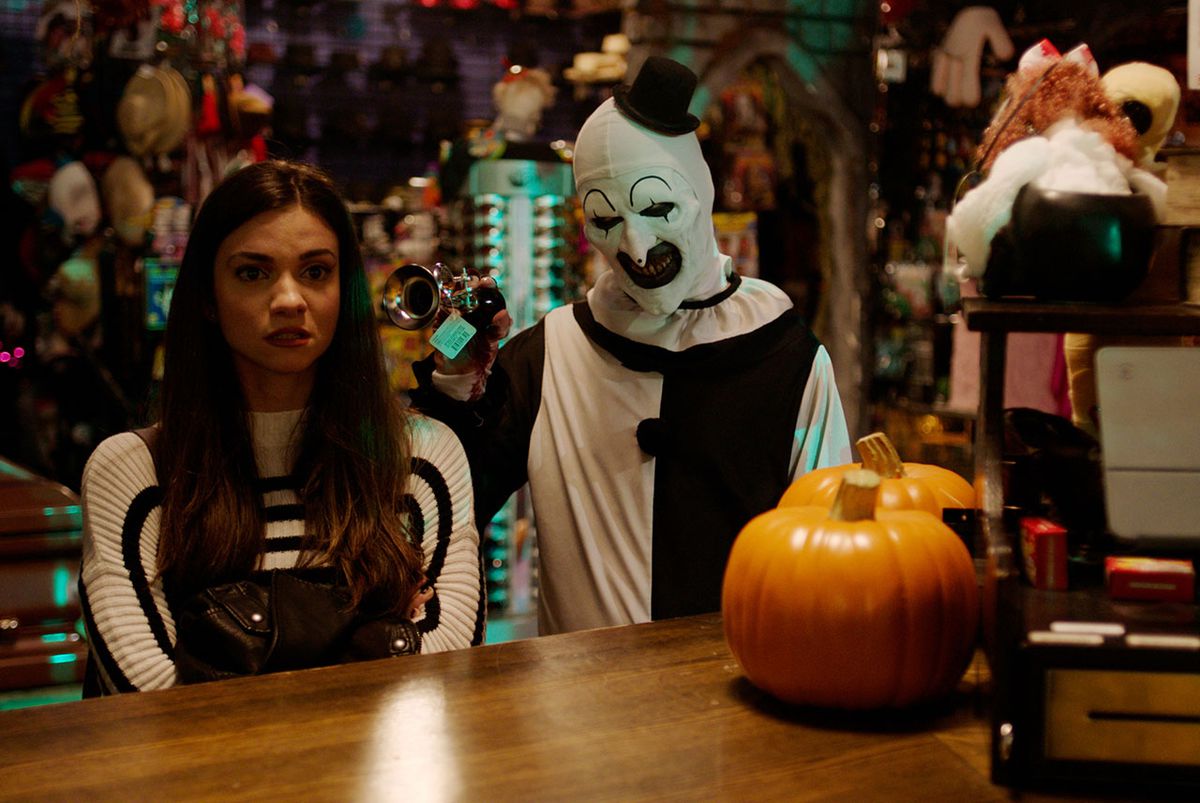 Une jeune femme debout au comptoir d'un magasin de décoration d'Halloween avec un homme lorgnant en maquillage de clown noir et blanc grinçant une corne dans son oreille.