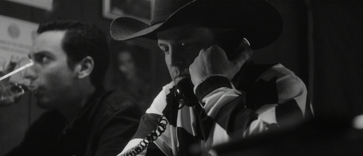 Un homme portant un chapeau de cow-boy tient un téléphone avec un cordon à son oreille dans une image en noir et blanc, avec une personne à côté de lui qui boit dans un verre.