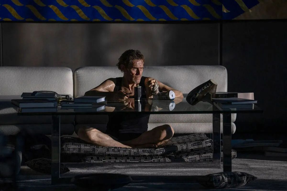Un homme nu et échevelé en caleçon (Willem Dafoe) est assis les jambes croisées devant une table en verre dans un salon sombre avec de la lumière provenant d'une fenêtre hors champ dans Inside.