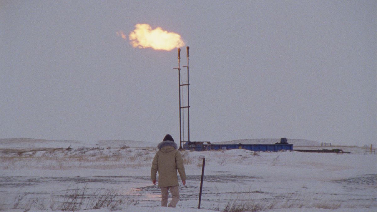 Un homme en parka marche dans la neige alors que les flammes jaillissent d'une cheminée devant lui dans How to Blow Up a Pipeline