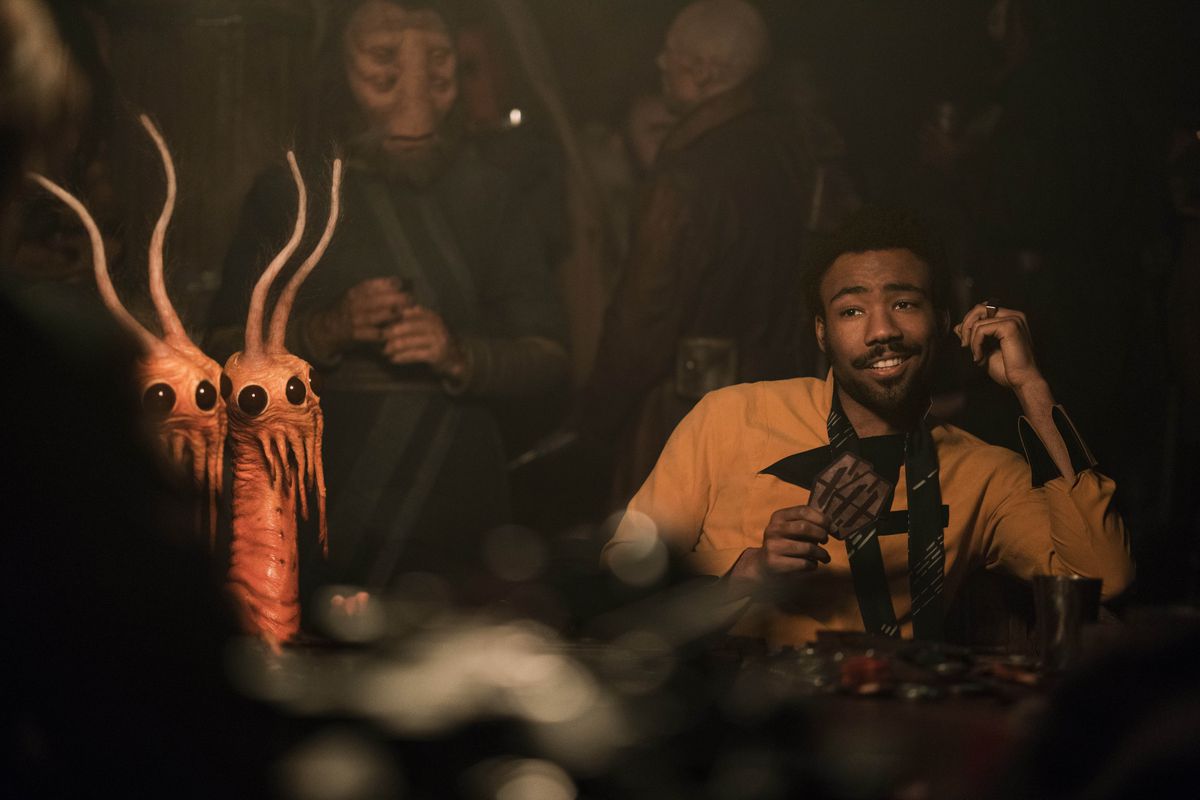 Donald Glover dans le rôle de Lando Calrissian dans Solo : Une histoire de Star Wars.  Il se prélasse discrètement à une table de jeu sabacc, entouré d'étranges extraterrestres. 