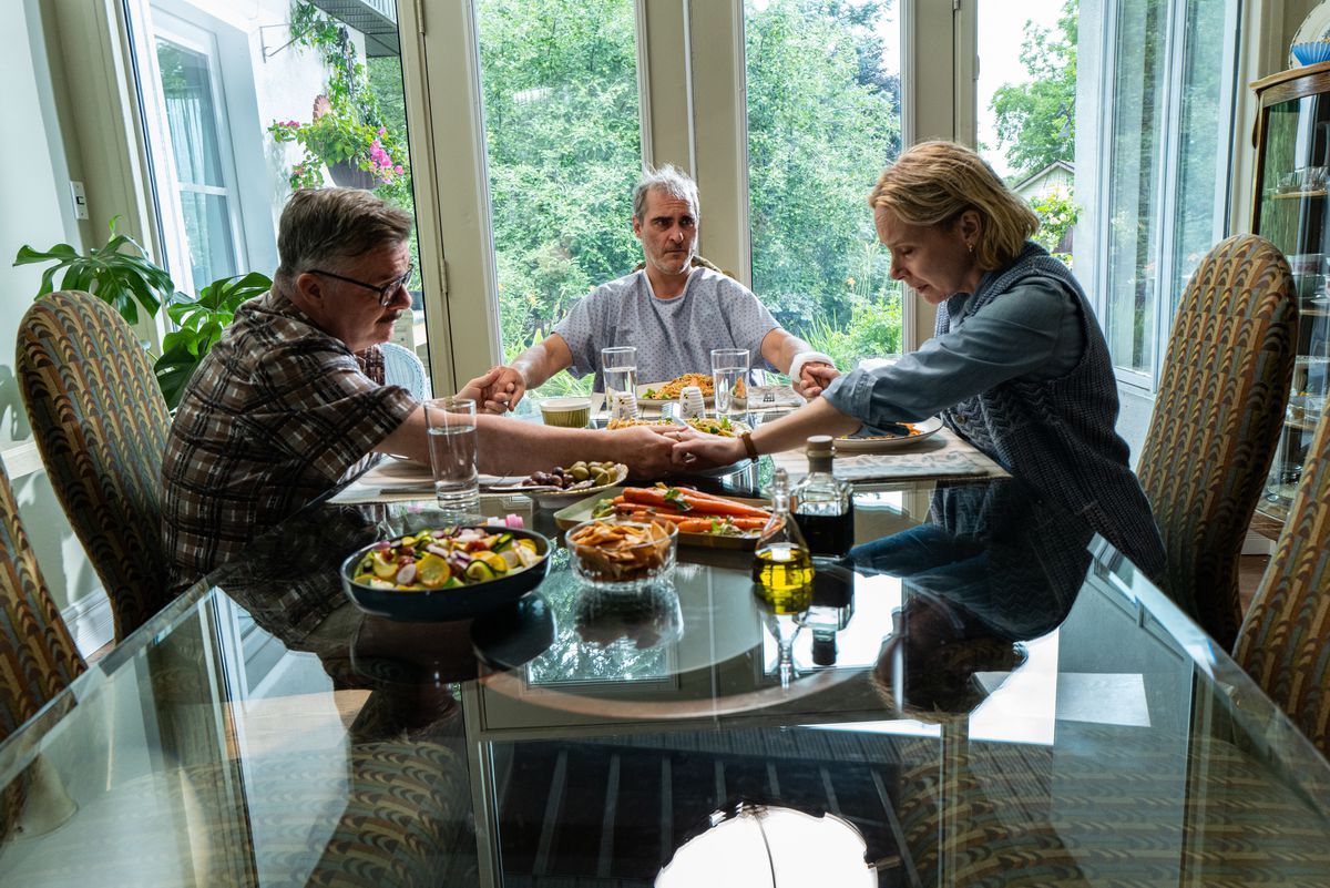 Nathan Lane, Joaquin Phoenix et Amy Ryan sont assis autour d'une table à dîner en se tenant la main et en priant dans Beau a peur