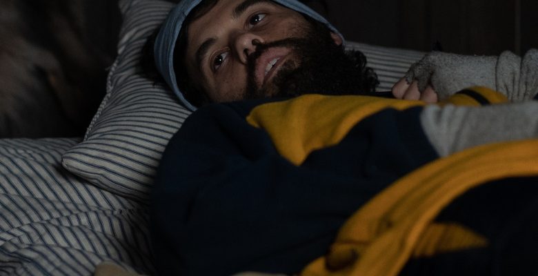 A man in yellow and blue winter athletic wear lays in bed with an empty expression; still from "Yellowjackets"
