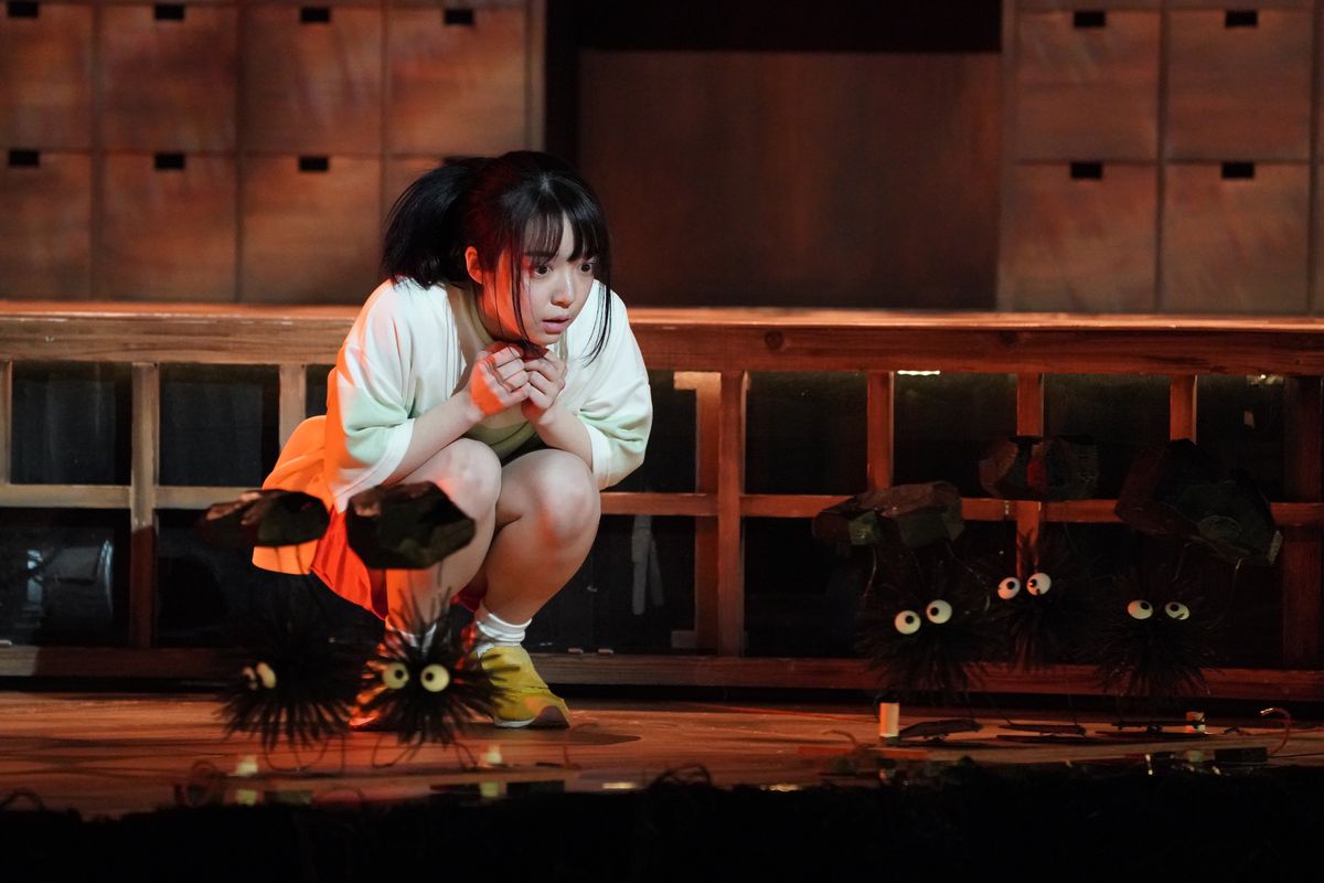 Chihiro s'accroupit et regarde les marionnettes de sprites de suie transportant du charbon se déplaçant sur la scène dans Spirited Away: Live on Stage