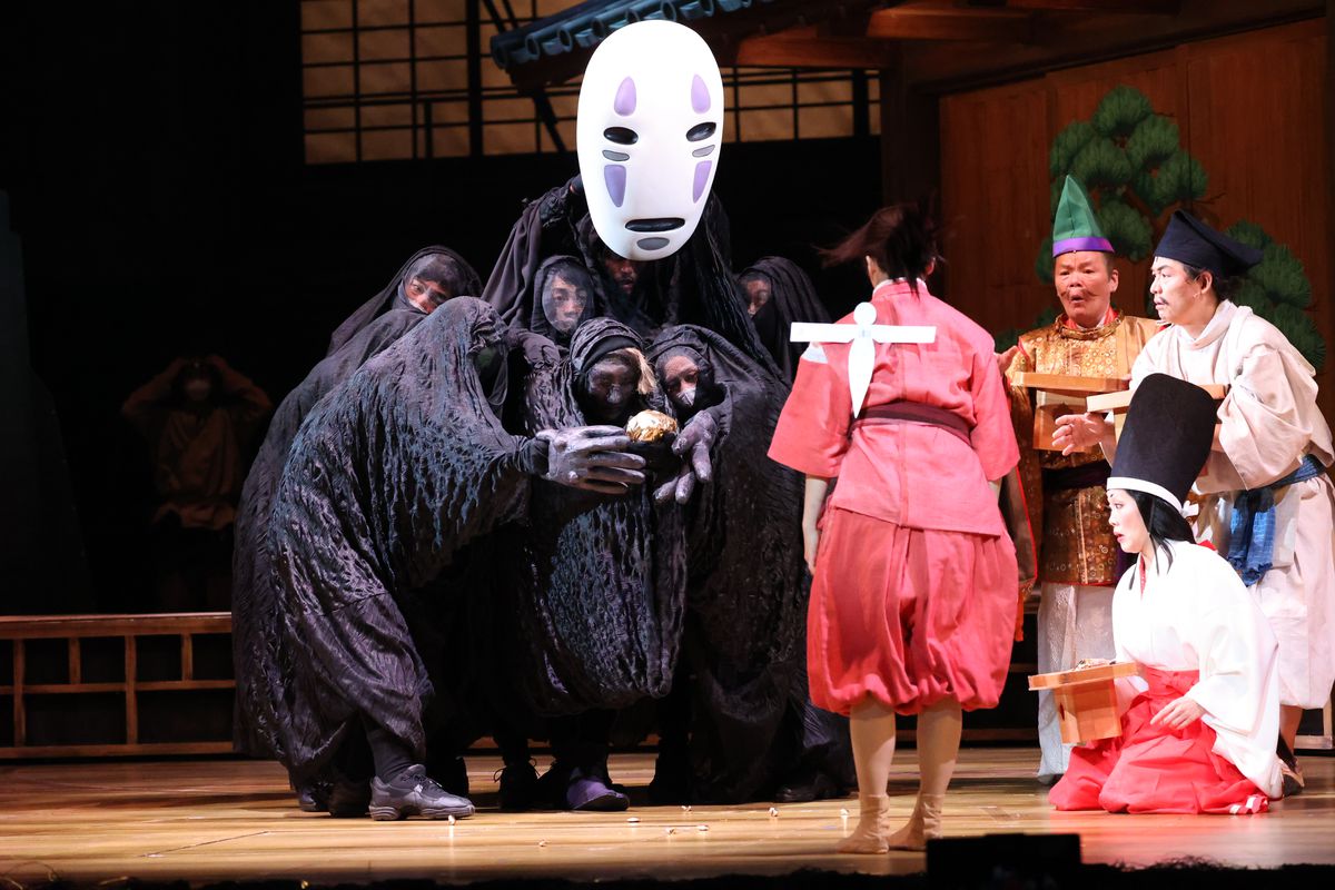 Chihiro (Mone Kamishiraishi, en robe de bain rose, avec un seul Shikigami blanc collé à son dos) fait face à Kaonashi (également connu sous le nom de No-Face, joué par un groupe d'artistes sous une bâche noire semi-transparente) comme d'autres employés du regard sur les bains de esprit dans Spirited Away: Live on Stage
