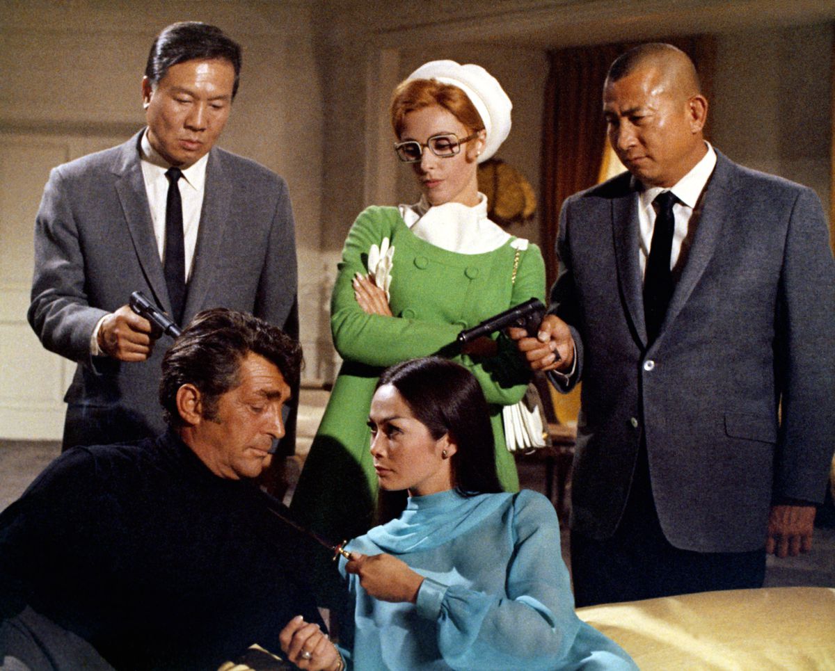Deux hommes de main asiatiques pointent des armes sur Matt Helm alors qu'une femme rousse avec des lunettes dans un manteau vert ricane derrière lui et qu'une femme asiatique en robe bleue tient un couteau