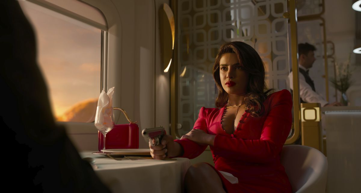 Priyanka Chopra Jonas dans le rôle de Nadia Sinh est assise à une table à manger dans un train alors que le soleil se couche par la fenêtre, illuminant sa robe rouge et l'arme qu'elle pointe sur un homme hors écran en face d'elle dans l'émission Prime Video Citadel.