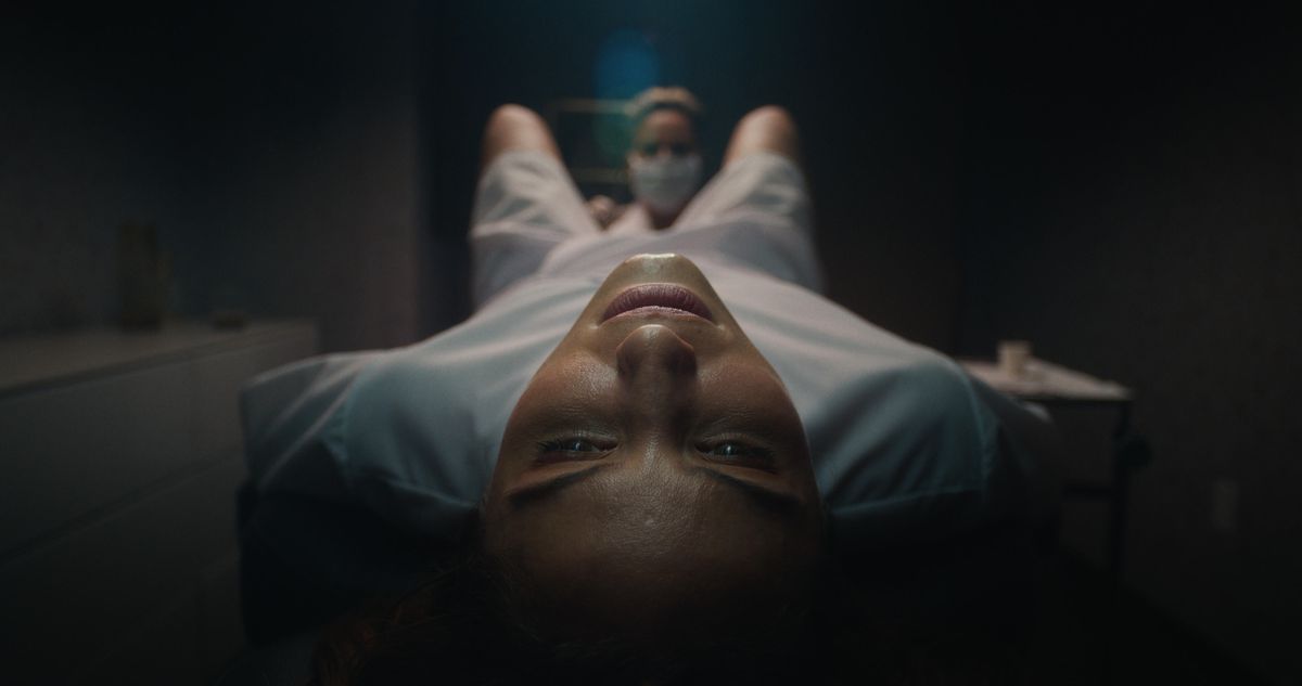 Un gros plan d'une femme (Dianna Agron) dans une chemise d'hôpital allongée sur une table pendant qu'une infirmière examine entre ses jambes dans Clock.