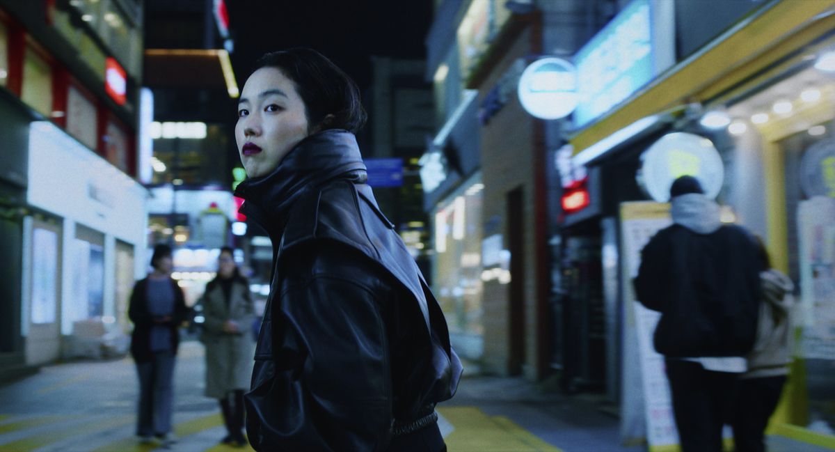 Jin-Min Park dans le rôle de Freddie, portant du rouge à lèvres rouge foncé dans un manteau de cuir noir, debout au milieu d'une rue de la ville coréenne la nuit dans Return to Seoul.