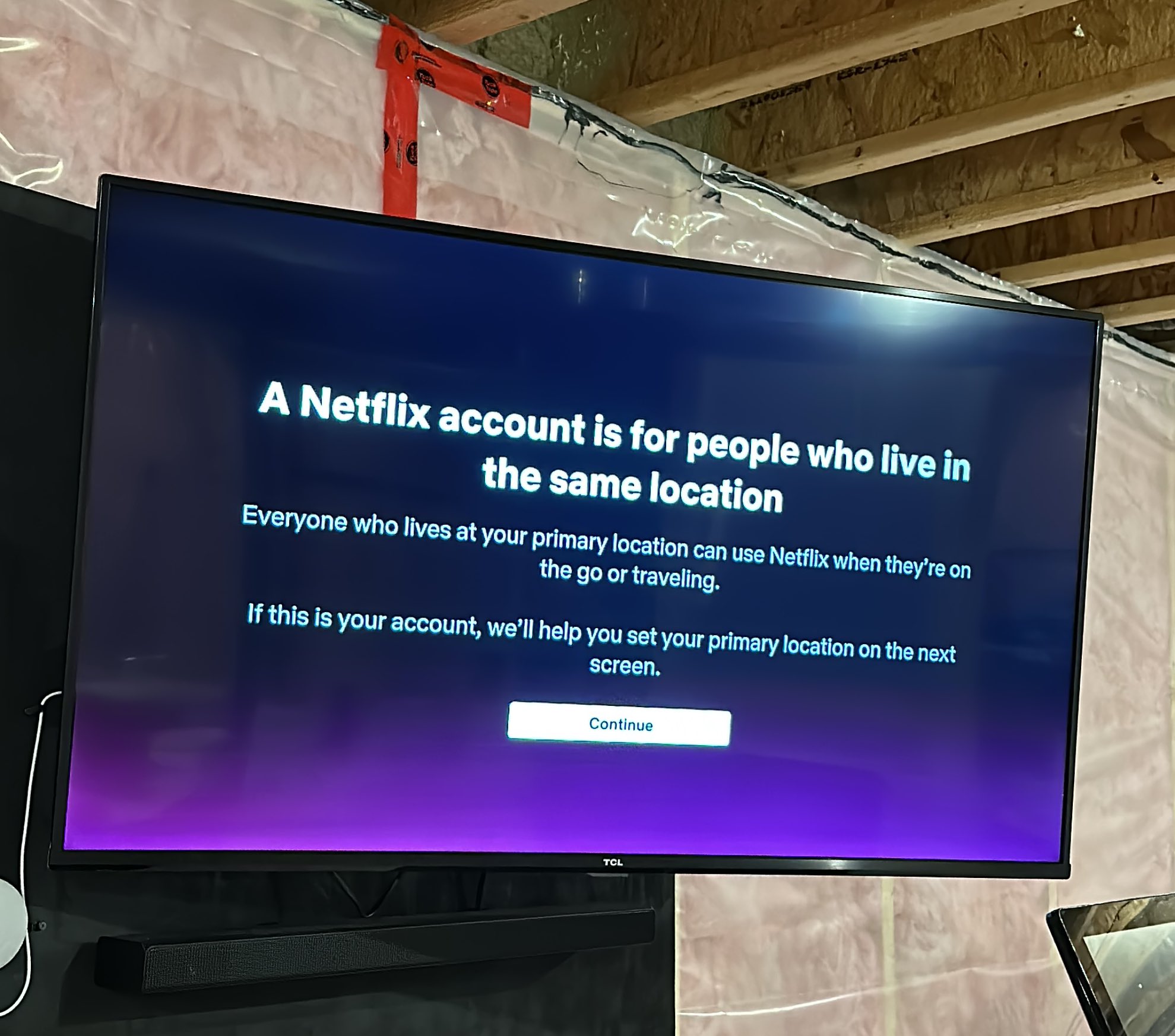 Un téléviseur monté sur un mur avec le message de répression du mot de passe Netflix : "Un compte Netflix est destiné aux personnes qui vivent au même endroit. Tous ceux qui vivent à votre emplacement principal peuvent utiliser Netflix lorsqu'ils sont en déplacement ou en voyage.  S'il s'agit de votre compte, nous vous aiderons à définir votre emplacement principal sur l'écran suivant." et un bouton Continuer.