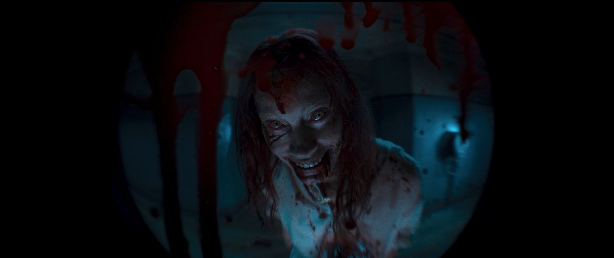 la Deadite Ellie donne un sourire monstrueux de l'autre côté d'un judas taché de sang dans Evil Dead Rise