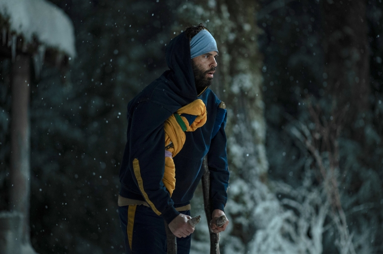 Un homme emmitouflé dans des vêtements d'hiver et sur des béquilles, debout à l'extérieur d'une cabane dans les bois la nuit avec de la neige au sol ;  encore de 