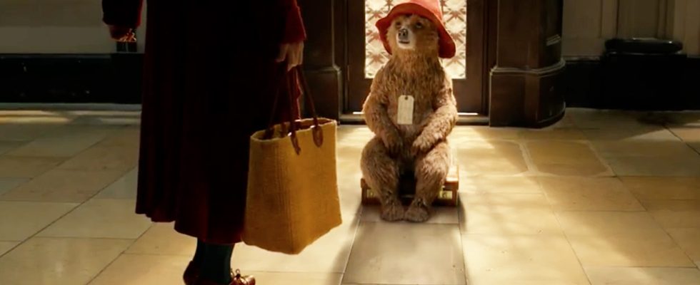 Paddington Bear sits on a suitcase; Mrs. Brown's legs appear in the foreground of the shot