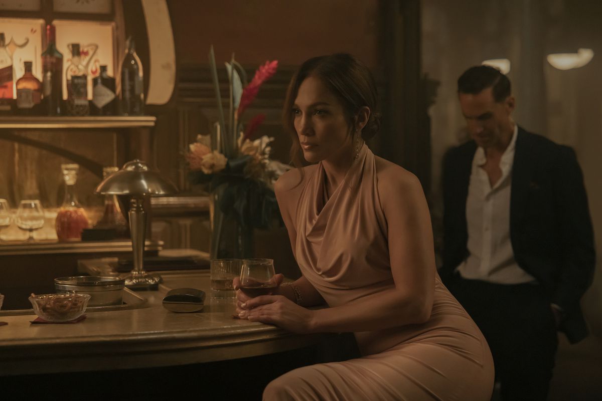 Jennifer Lopez est assise dans un bar avec un verre de vin tout en portant une jolie robe dans The Mother.  Joseph Fiennes se profile derrière elle, vêtu d'un costume.