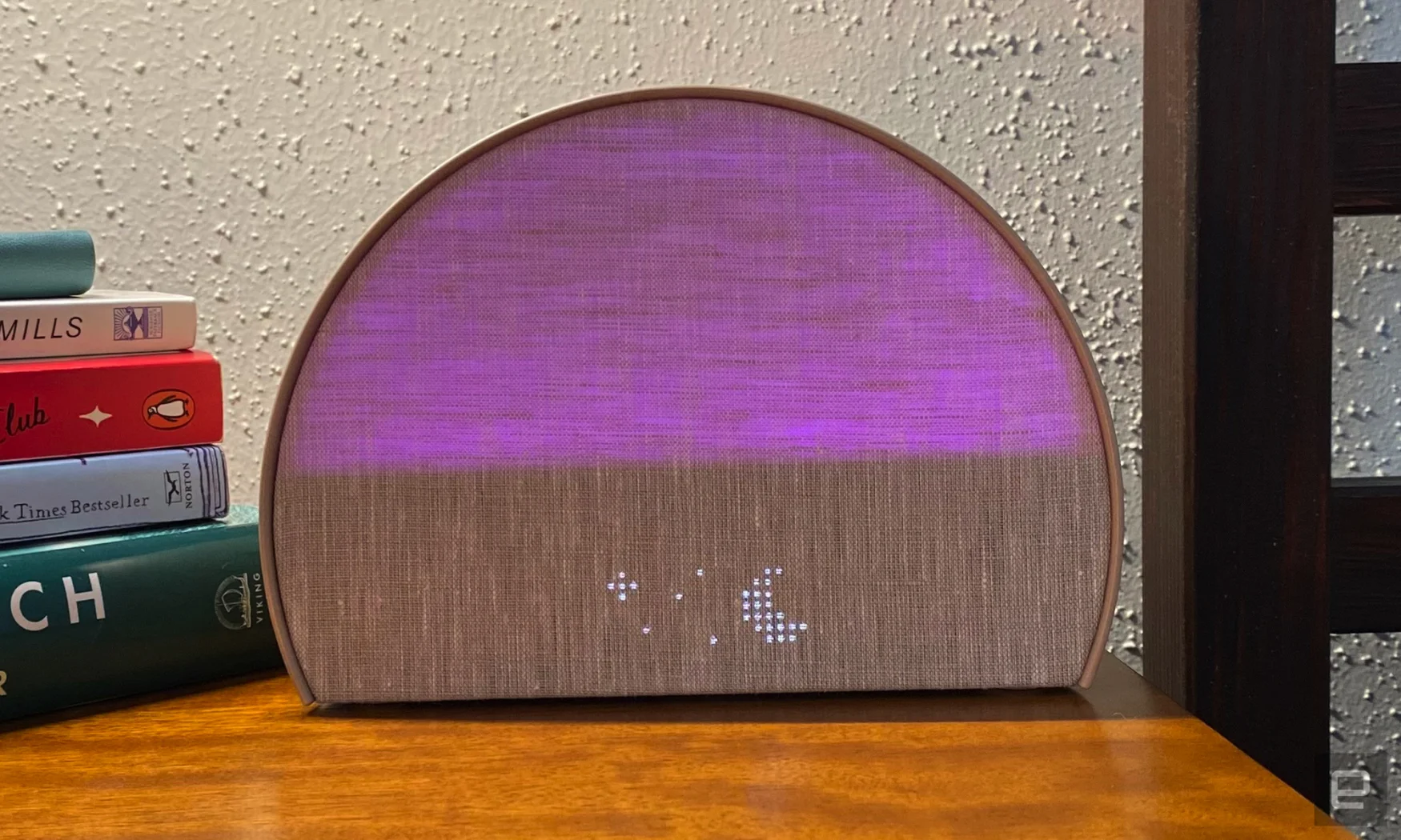 Le Hatch Restore 2 s'allume en violet et affiche une lune et des étoiles à matrice de points.  L'unité repose sur une table de chevet en bois et le coin de quelques livres sont empilés en arrière-plan. 