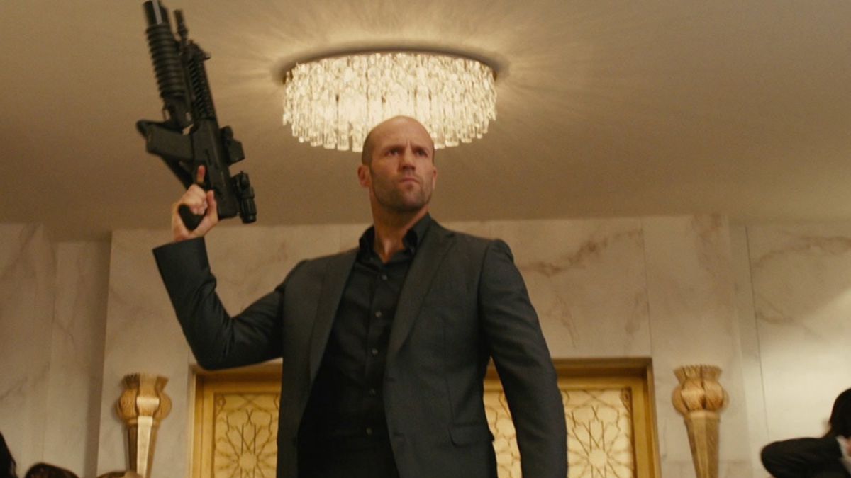 Jason Statham pointe un fusil d'assaut en l'air devant un lustre dans Furious 7.
