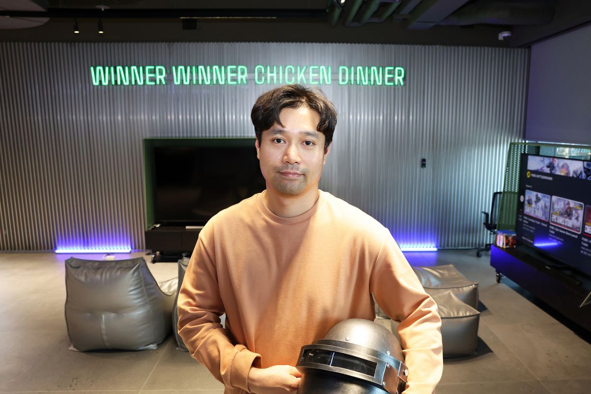 Une photo de Taeyhun Kim de PUBG Studios tenant le casque PUBG classique et se tenant devant une enseigne au néon indiquant «Winner winner chicken dinner».