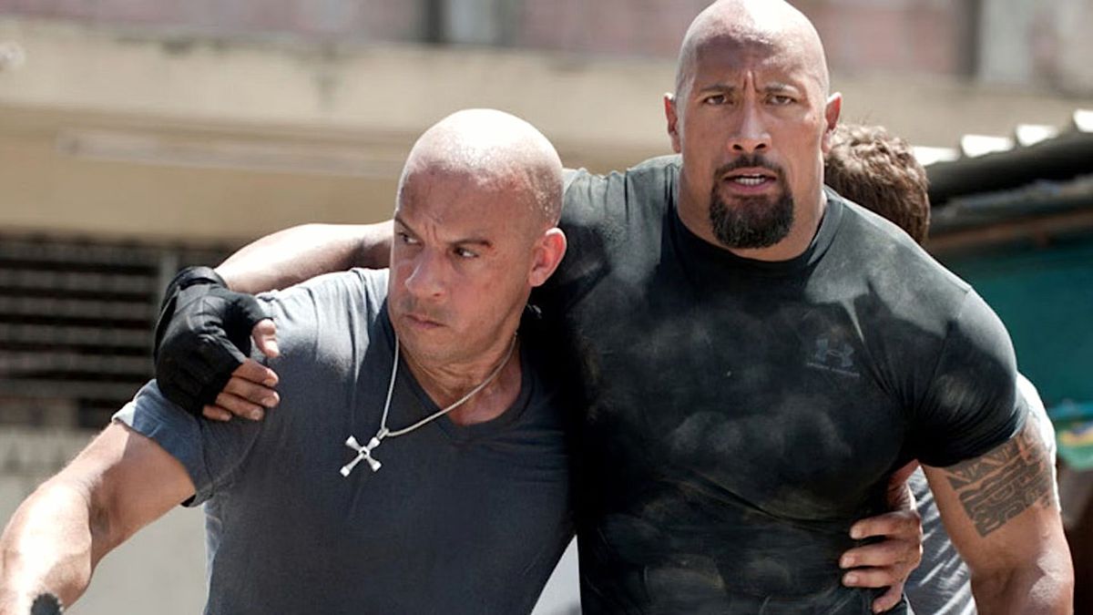 Dom (Vin Diesel) et Hobbs (Dwayne Johnson) se soutiennent mutuellement avec leurs bras autour de la taille dans Fast Five.  Le crucifix de Dom, qui n'est actuellement pas remis à un membre de sa famille pour être gardé en lieu sûr, est bien en vue autour de son cou.