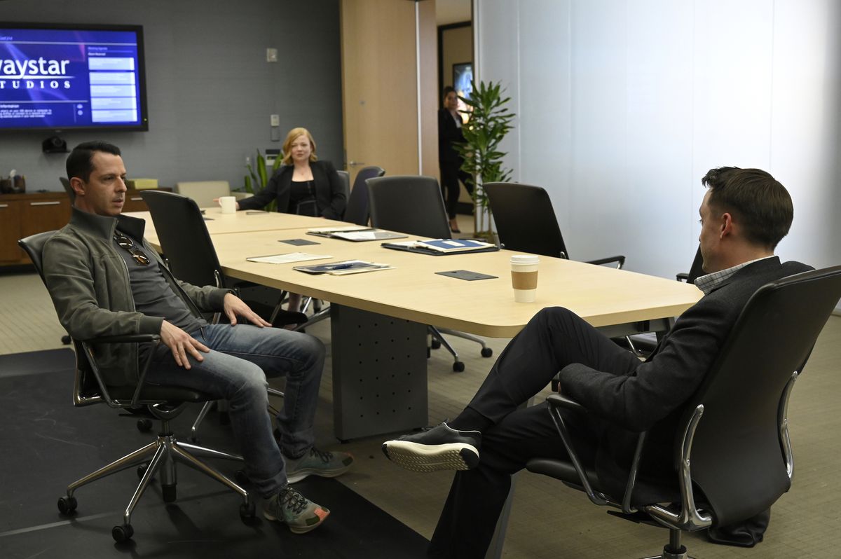 Shiv (Sarah Snook) est assise de l'autre côté à l'arrière-plan à une table de conférence tandis que ses frères Kendall (Jeremy Strong) et Roman (Kieran Culkin) s'assoient et parlent plus près du premier plan