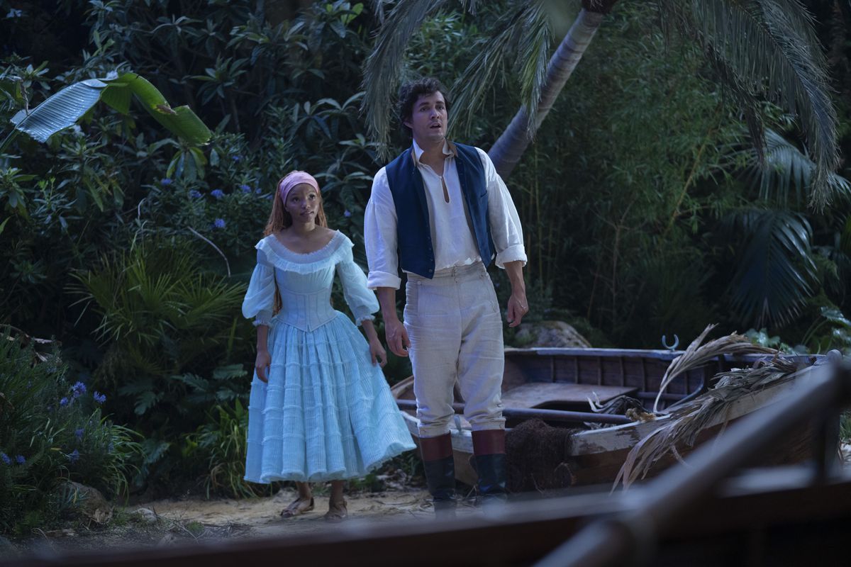 Eric et Ariel dans le live-action Little Mermaid se tiennent sur une plage.  Ariel porte une robe bleu clair, tandis qu'Eric porte une chemise blanche avec un gilet bleu et un pantalon kaki.