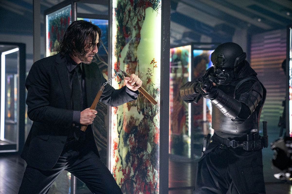 Keanu Reeves dans le rôle de John Wick tenant des num-chuks derrière un panneau de verre se préparant à tendre une embuscade à un soldat en armure de samouraï noir dans John Wick : Chapitre 4.