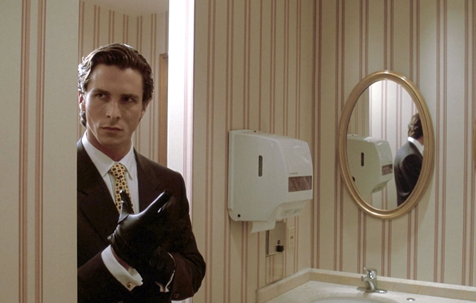 Patrick Bateman (Christian Bale) entre dans une salle de bain en enfilant un gant noir dans le film American Psycho.