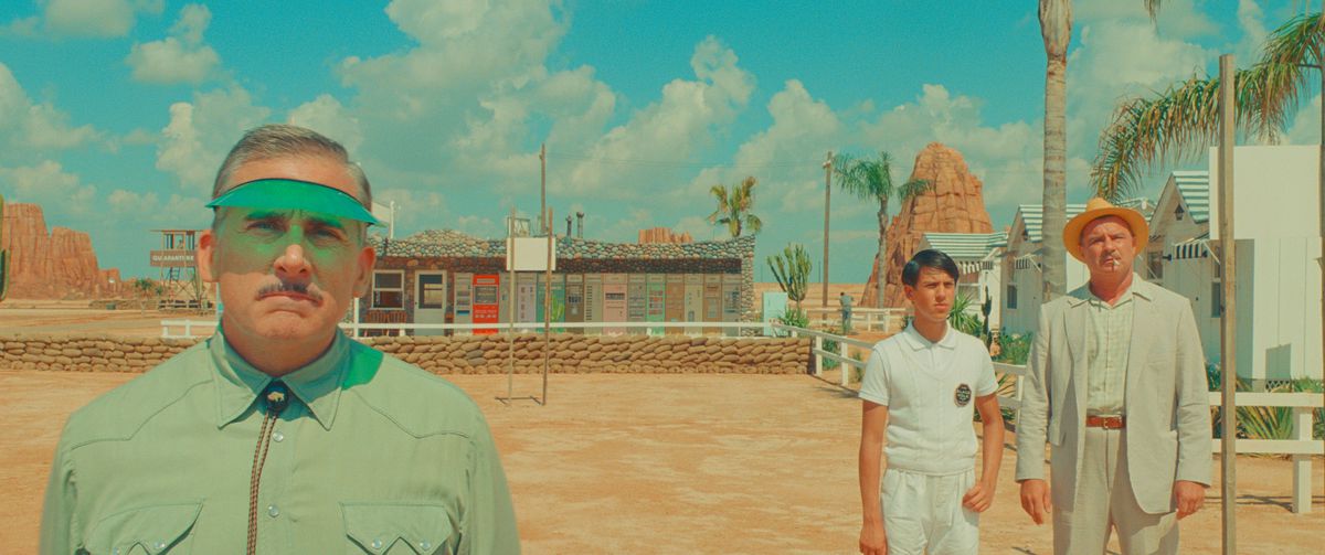 Un directeur de motel (Steve Carell) se tient devant un motel du désert, portant un parasol vert et regardant la caméra, tandis que deux personnes en vêtements pâles (Aristou Meehan et Liev Schreiber) se tiennent au loin derrière lui dans Asteroid City de Wes Anderson