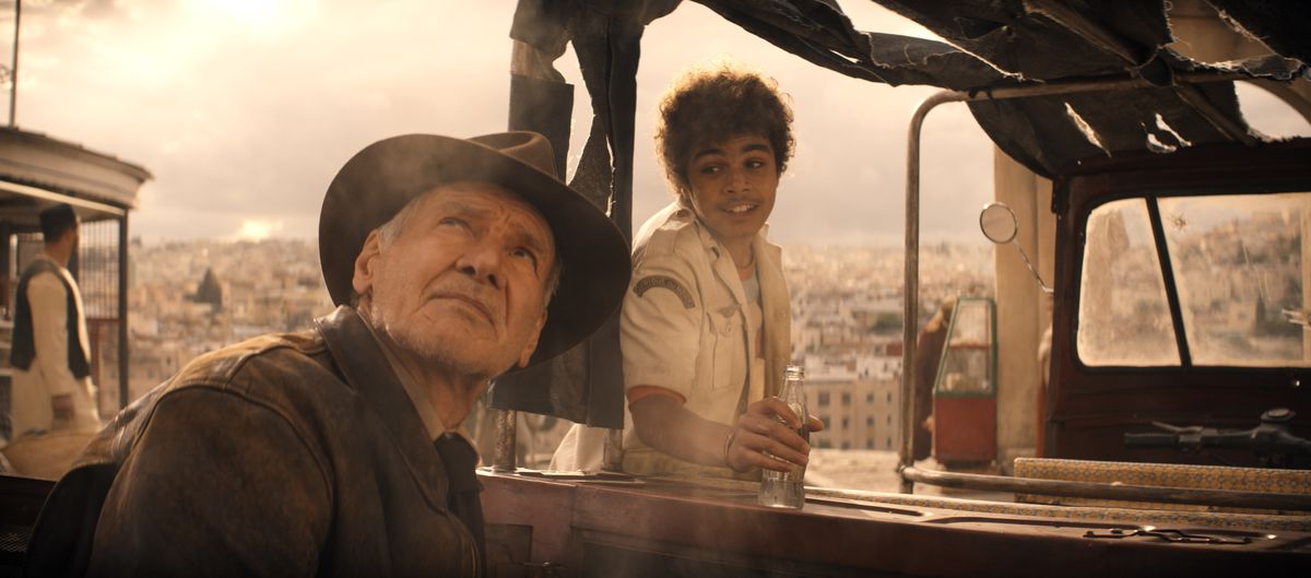 Indiana Jones (Harrison Ford) lève les yeux vers un ciel gris couvert tandis qu'un adolescent souriant (Ethann Isidore) essaie de lui parler alors qu'il se prépare pour un tour dans son camion dans Indiana Jones et le cadran du destin