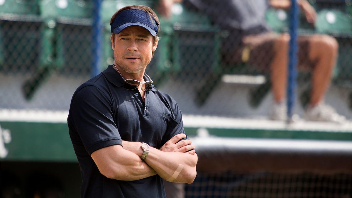 Un homme en polo bleu marine (Brad Pitt) portant un chapeau bleu se tenant debout sur le côté d'un terrain de baseball.