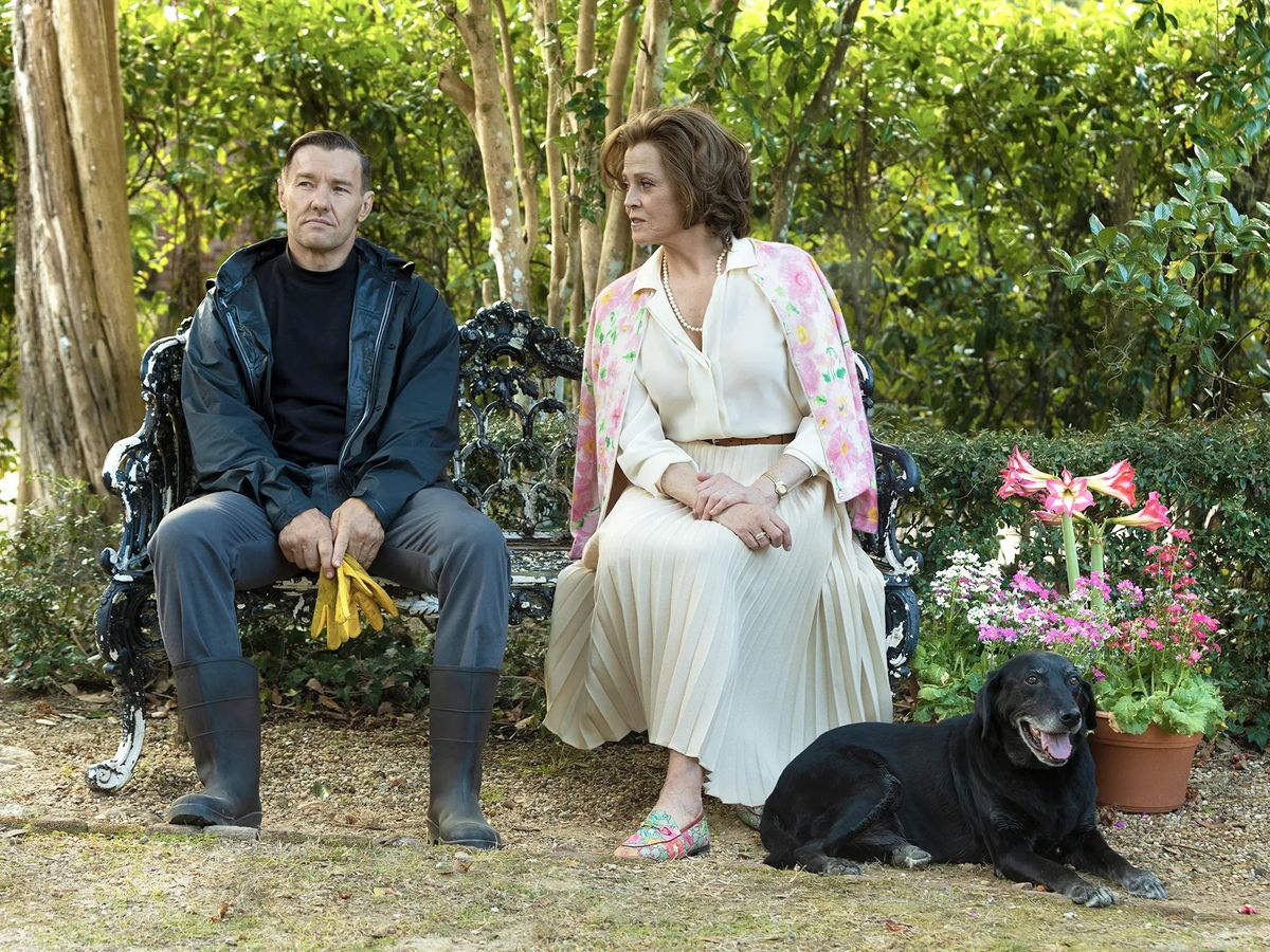 (LR) Joel Edgerton et Sigourney Weaver assis sur un banc près d'un arrangement de fleurs en pot et d'un chien noir dans Master Gardener.