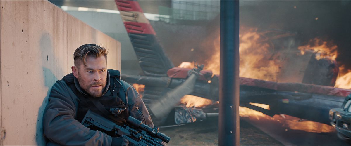 Le mercenaire Tyler Rake (Chris Hemsworth) s'accroupit contre un mur de béton et tient un fusil de sniper lourd alors que l'épave d'un hélicoptère brûle à côté de lui dans Extraction 2