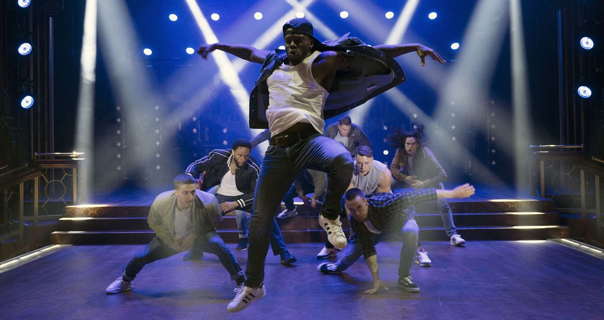 Un danseur noir en jeans et une casquette de baseball à l'envers saute haut dans les airs tandis que d'autres danseurs masculins s'accroupissent derrière lui sur une scène éclairée en bleu sillonnée de projecteurs blancs brillants dans une scène de Magic Mike's Last Dance