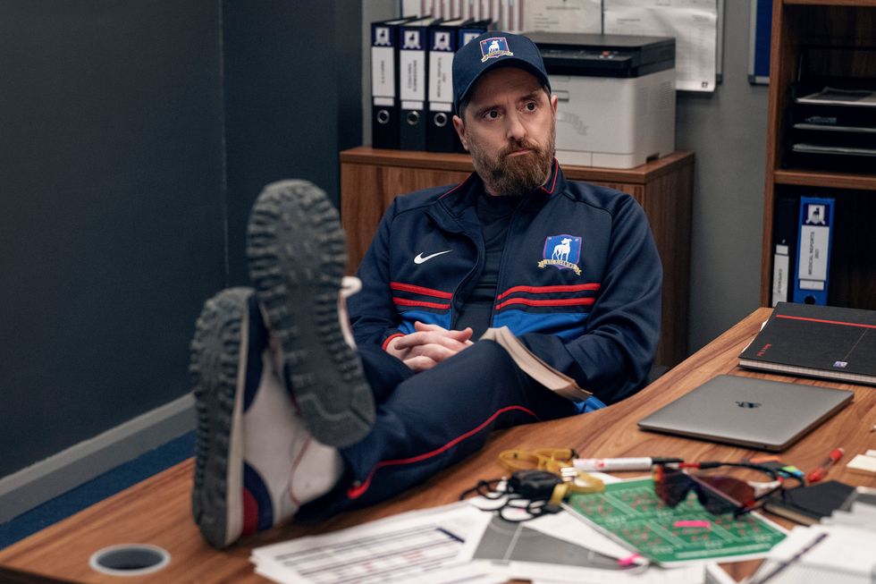 dans une scène de la saison 3 de ted lasso, l'entraîneur beard est assis les pieds sur le bureau du bureau de l'équipe