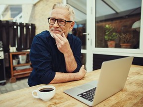 Un homme plus âgé assis devant un ordinateur portable et une tasse de café sur une terrasse à l'extérieur.