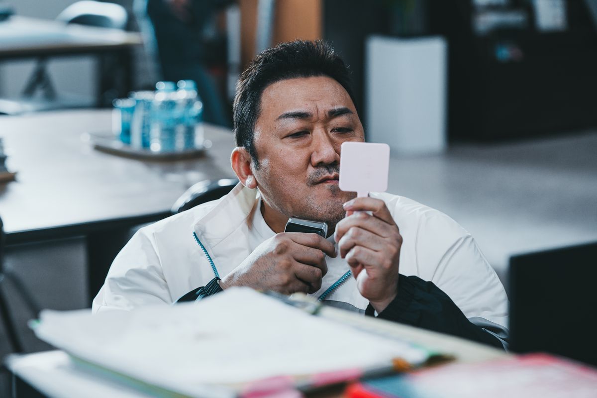 Ma Dong-seok tient un miroir à main tout en se rasant le visage à son bureau dans The Roundup: No Way Out.