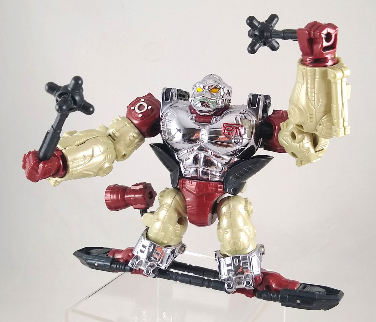 Une photo d'un jouet Transformers exclusif à la convention Apelinq, un singe en métal rouge, noir, argent et crème debout sur un hoverboard noir et brandissant des masses noires