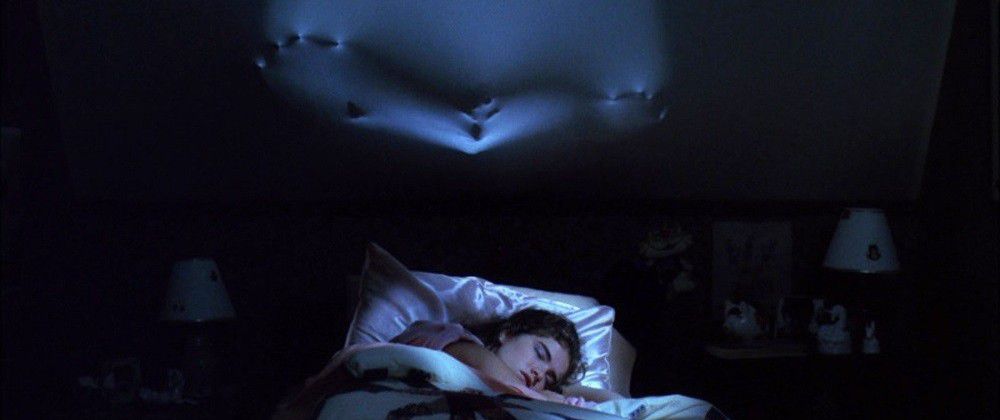 Une femme dormant profondément dans son lit alors qu'un spectre macabre pousse visiblement contre le plafond au-dessus d'elle dans A Nightmare on Elm Street.